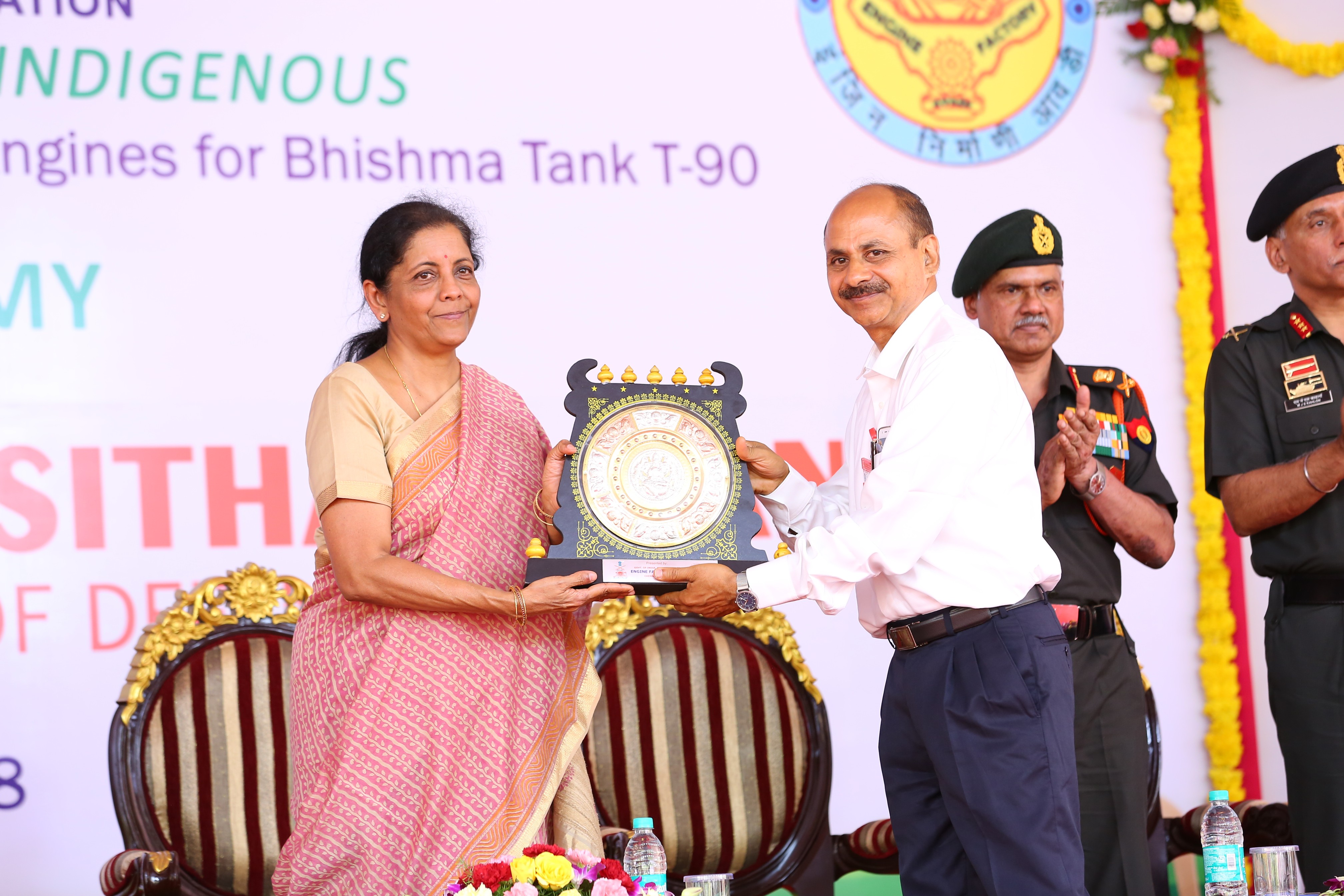 Nirmala Sitaraman in a pink saree and a man in a white shirt present an award plaque. Two men in military uniforms stand in the background, clapping.